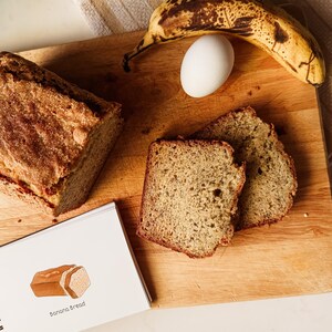 Könnte beinhalten: Ein Holzbrett mit einem Laib Bananenbrot, zwei Scheiben Bananenbrot, einer Banane und einem Ei. Ein Notizbuch mit den Worten "Banana Bread" liegt auf dem Brett.