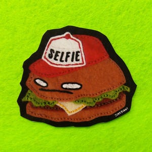 May include: A felt burger patch with a red and white hat that says "SELFIE". The burger has a face and is wearing a hat. The patch is on a green background.