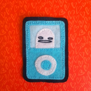 以下が含まれることがあります： 青いフェルト生地に、黒い口と目をつけた小さな白い漫画のキャラクターが、青いフェルト生地の四角形の中に描かれています。四角形の底の中央には、白いフェルト生地の円が描かれています。四角形は黒いフェルト生地の縁で囲まれています。