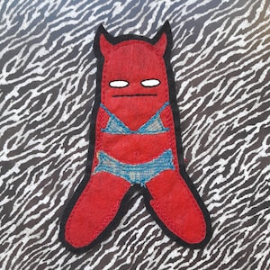 May include: Red felt appliqué of a devil wearing a blue bikini. The devil has horns and a serious expression. The appliqué is on a black and white zebra print background.
