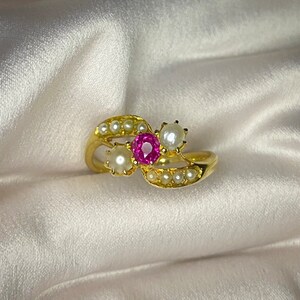 May include: A gold ring with a pink gemstone and three white pearls. The ring is set on a white satin background.