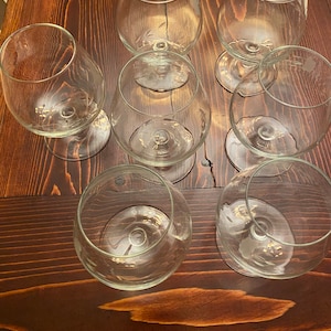 May include: A collection of seven clear brandy snifters with etched floral designs. The glasses are arranged on a dark brown wooden surface, showcasing their elegant shape and delicate details. The stems are short and the bowls are round.