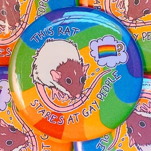 May include: A collection of colorful pinback buttons. The central button features a cartoon rat with the text "THIS RAT STARES AT GAY PEOPLE" in a rainbow design. Other buttons with similar designs are visible.