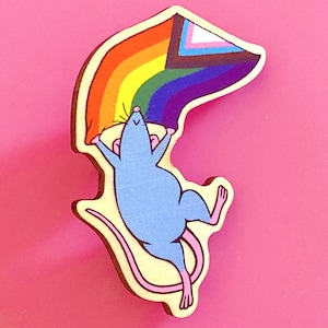 Puede incluir: Un pin de madera con un ratón de dibujos animados azul claro sosteniendo una bandera del Orgullo arcoíris. El pin tiene un borde amarillo claro y está sobre un fondo rosa. La bandera incluye los colores de la bandera del Orgullo, así como los colores de la bandera del Progreso del Orgullo.