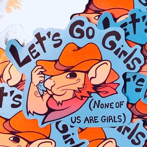 Puede incluir: Una pegatina con un ratón de dibujos animados con un sombrero de vaquero y una bufanda roja. La pegatina tiene el texto "Let's Go Girls" y "(None of us are girls)" en negro sobre un fondo azul claro. El ratón es marrón y naranja.