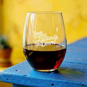 May include: A stemless wine glass filled with red wine, set against a blue wooden surface and a yellow background. The glass has white text that reads "Don't let the Bastards Get You Down!"