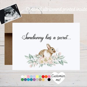 May include: A white card with the text "Somebunny has a secret..." and a watercolor illustration of a brown bunny sitting in a bed of pink flowers and green leaves. The card is surrounded by a brown envelope.