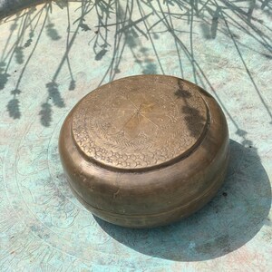 May include: A round, antique brass container with an etched floral design on the lid. The container is sitting on a teal-coloured surface with a floral pattern. The container is in the sunlight, casting a shadow.