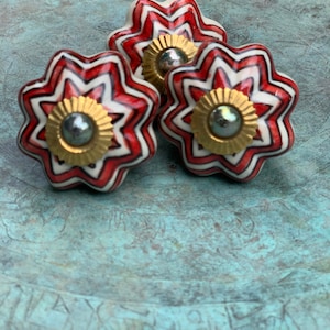May include: Three ceramic drawer knobs with a starburst design. The knobs are white with red and black stripes and have a gold centre with a silver button.