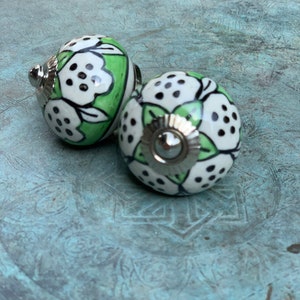 May include: Two ceramic knobs with a white and green floral pattern and black dots. The knobs have a silver metal base.
