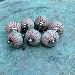 May include: Eight ceramic knobs with a white base and red accents. The knobs have a textured surface and a silver metal base.
