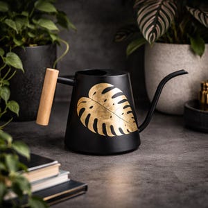 Könnte beinhalten: Schwarze Gießkanne mit langem Ausguss und Holzgriff. Auf der Seite der Kanne befindet sich ein goldenes Blattdesign. Die Gießkanne steht auf einer dunkelgrauen Oberfläche, mit Pflanzen im Hintergrund.