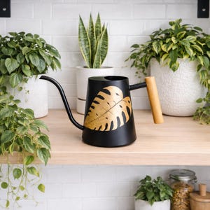 Könnte beinhalten: Schwarze Metall-Gießkanne mit langem, gebogenem Ausguss und Holzgriff. Auf der Seite ist ein goldenes Blattmuster abgebildet. Die Gießkanne steht auf einem Holzregal mit verschiedenen Topfpflanzen im Hintergrund.