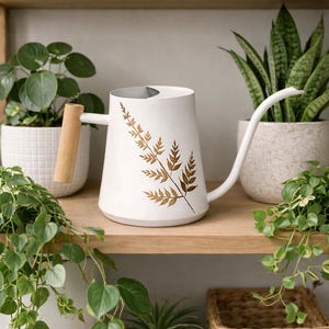 Könnte beinhalten: Weiße Gießkanne mit langem Ausguss und Holzgriff. Die Kanne hat ein goldenes Farn-Design. Topfpflanzen stehen auf Holzregalen. Ein Weidenkorb ist ebenfalls sichtbar.