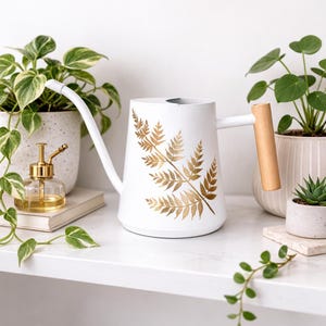Könnte beinhalten: Weiße Gießkanne aus Metall mit langem Ausguss und Holzgriff. Die Kanne hat ein goldenes Farnblatt-Design. Die Gießkanne ist von Topfpflanzen und einer kleinen goldenen Sprühflasche umgeben.