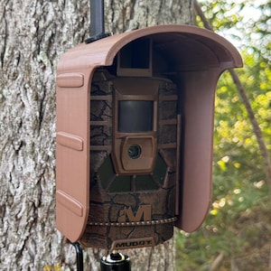 May include: A brown camouflage trail camera with a black antenna and a brown protective cover. The camera is mounted on a black metal tree mount. The camera has a black lens and a black sensor. The camera is labeled "Muddy".
