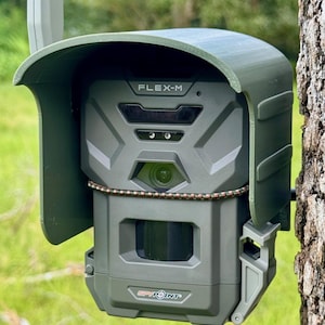 May include: A green trail camera with a camouflage pattern and a black lens. The camera has a black and white LCD screen and the text "FLEX-M" and "SPYPOINT" on it. The camera is mounted on a tree.