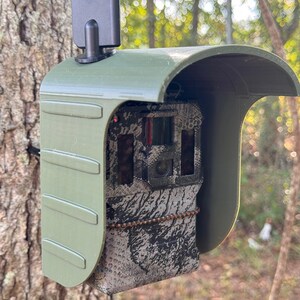 May include: A green camouflage trail camera with a black antenna attached to the top. The camera is mounted on a tree trunk.