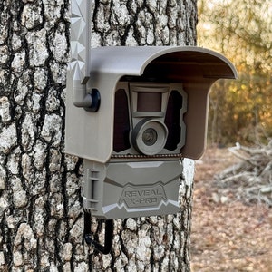 May include: A brown and gray wildlife camera with a black lens and a camouflage pattern. The camera is mounted on a tree and has a black antenna. The camera has a black protective cover and the text "REVEAL X-PRO" is printed on the front.