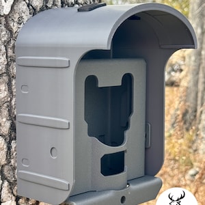 May include: A gray trail camera enclosure mounted on a tree. The enclosure has a curved top and a hinged front panel. The Sahara Outdoors logo is visible in the lower right corner, and a Facebook logo is in the lower left.