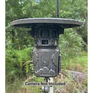 May include: A dark gray trail camera with the text "PRO 2.0" and "STEALTH CAM". The camera is mounted on a metal pole and has a circular top. The text "Camera Not Included" is at the bottom.