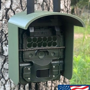 May include: A green weatherproof camera housing with a black strap and a black trail camera mounted inside. The camera has a black lens and a black and gray display. The housing is mounted to a tree trunk. The text "MADE IN USA" is visible in the bottom right corner of the image.