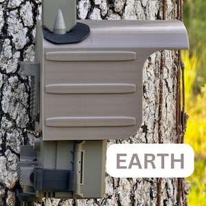 May include: A gray wildlife camera with a camouflage rain shield attached to a tree trunk. The camera has a black strap and a black and gray lens. The text "EARTH" is visible on the tree trunk.