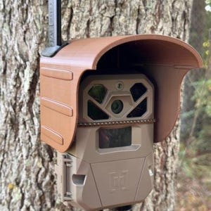 May include: A brown camouflage trail camera with a brown protective cover mounted to a tree. The camera has a black lens and a black antenna.