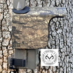May include: A camouflage-patterned trail camera mounted on a tree trunk. The camera has a Realtree Officially Licensed Products logo. The camera is designed to blend in with the natural environment.
