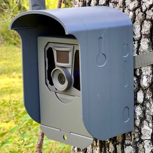 May include: A gray wildlife camera with a black lens is mounted to a tree trunk. The camera is protected by a gray weatherproof housing with a curved top.