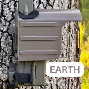 May include: A tan-colored camera housing mounted on a tree trunk. The housing has a protective cover with three horizontal ridges and a pointed top. The word "EARTH" is displayed in a white rounded rectangle.