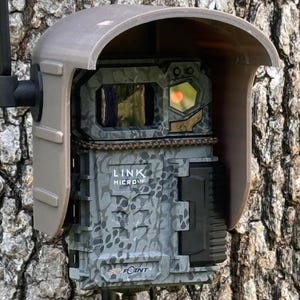 May include: A gray and black wildlife camera with a camouflage pattern. The camera has a black lens cover and a gray protective cover. The camera is mounted on a tree and has the text "LINK MICRO" and "SPOT POINT" printed on it.