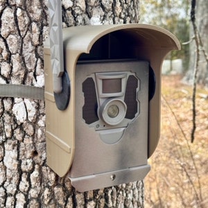 May include: A brown trail camera with a black lens and a camouflage pattern is mounted to a tree trunk. The camera has a black antenna and a brown rain shield.