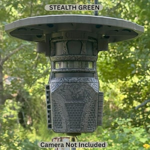 May include: Stealth green camera mount with a circular top and a textured body. The mount has the word "ORBIT" on it. The text "Camera Not Included" is at the bottom. The mount is designed to hold a camera.