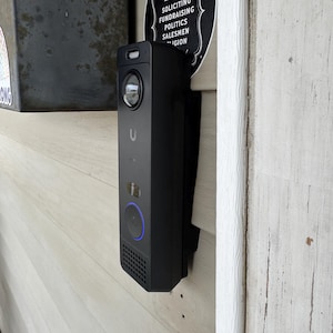 May include: A black smart doorbell with a camera and a blue illuminated ring. The doorbell is mounted on a light-colored wooden wall next to a sign that reads "NO SOLICITING FUNDRAISING POLITICS SALESMEN RELIGION."