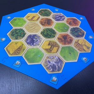 Op de afbeelding: Een blauw zeshoekig spelbord met 19 zeshoekige tegels. De tegels zijn in verschillende kleuren en tonen afbeeldingen van velden, bossen, bergen en schapen. Het bord is omgeven door een blauwe rand.