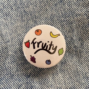 Może przedstawiać: Biały guzik z kolorowym wzorem owoców i napisem "fruity" napisanym czarnym kursywą. Guzik jest przypięty do niebieskiego materiału dżinsowego.