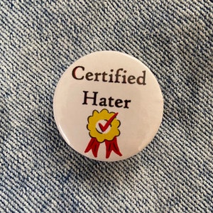 May include: White button with the text "Certified Hater" and a yellow checkmark with a red ribbon.