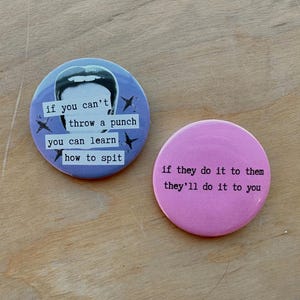 Op de afbeelding: Twee ronde buttons. De linker button is lichtblauw met een zwart-witte afbeelding van een mond en de tekst: If you can't throw a punch you can learn how to spit. De rechter button is effen roze met de tekst: if they do it to them they'll do it to you.