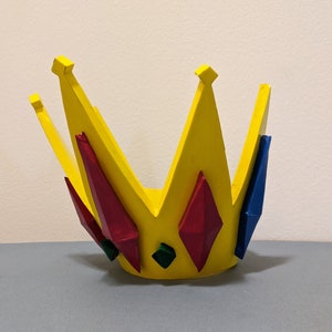 May include: A yellow crown with red, blue, and green gems. The crown is made of foam and is painted yellow. The gems are painted red, blue, and green.