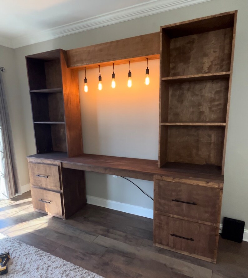 K&ouml;nnte beinhalten: Ein ma&szlig;gefertigter Holzschreibtisch mit integrierten Regalen und Schubladen. Der Schreibtisch hat eine dunkelbraune Oberfl&auml;che, mehrere Regale, zwei Schubladens&auml;tze und eine eingebaute Leuchte mit f&uuml;nf h&auml;ngenden Gl&uuml;hbirnen. Ideal f&uuml;r ein Homeoffice oder Arbeitszimmer.