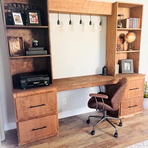 K&ouml;nnte beinhalten: Ein gro&szlig;er Holzschreibtisch mit integrierten Regalen und Schubladen. Der Schreibtisch hat einen braunen B&uuml;rostuhl aus Leder und h&auml;ngende Gl&uuml;hbirnen. Die Regale beherbergen B&uuml;cher, gerahmte Fotos und dekorative Gegenst&auml;nde.