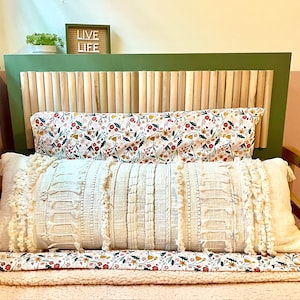 Op de afbeelding: Een bed met een groene hoofdeinde en een ontwerp van houten latten. Het bed is gemaakt met een wit en bloemig patroon dekbedovertrek en een wit kussen met franjes. De hoofdeinde heeft een bord met de tekst "Live Life".