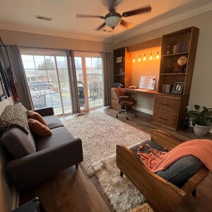 K&ouml;nnte beinhalten: Ein Wohnzimmer mit einem dunkelbraunen Sofa, einem Holzstuhl und einem Schreibtisch mit eingebautem B&uuml;cherregal. Ein flauschiger Teppich bedeckt den Boden. Der Raum hat einen Deckenventilator und eine Schiebet&uuml;r.