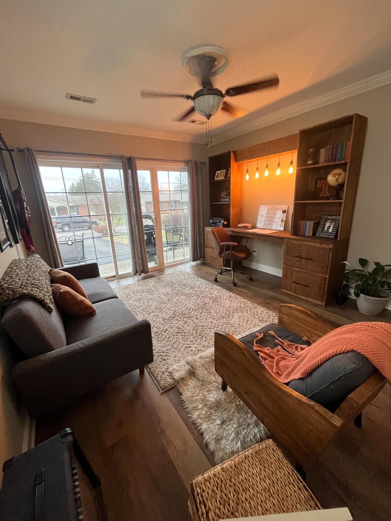 K&ouml;nnte beinhalten: Ein Wohnzimmer mit einem dunkelbraunen Sofa, einem Holzstuhl und einem Schreibtisch mit eingebautem B&uuml;cherregal. Ein flauschiger Teppich bedeckt den Boden. Der Raum hat einen Deckenventilator und eine Schiebet&uuml;r.