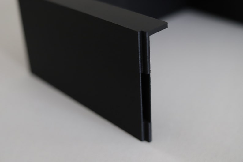 May include: Close-up of a black plastic corner piece with a smooth, matte finish. The corner piece has a slight curve and a small lip on the top edge.