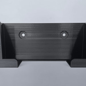 May include: A black plastic wall mount with two holes for screws. The mount has two vertical slots on the sides for holding a device.