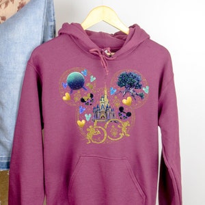 May include: Burgundy hoodie with a gold Mickey Mouse ears design featuring the Cinderella Castle, a Spaceship Earth, and a Tree of Life. The design also includes the number 50 in gold.