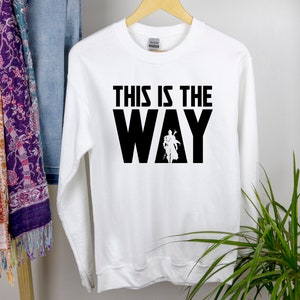 May include: White long-sleeved sweatshirt with black text that reads "This is the Way" and a silhouette of a person in the middle of the word "Way".