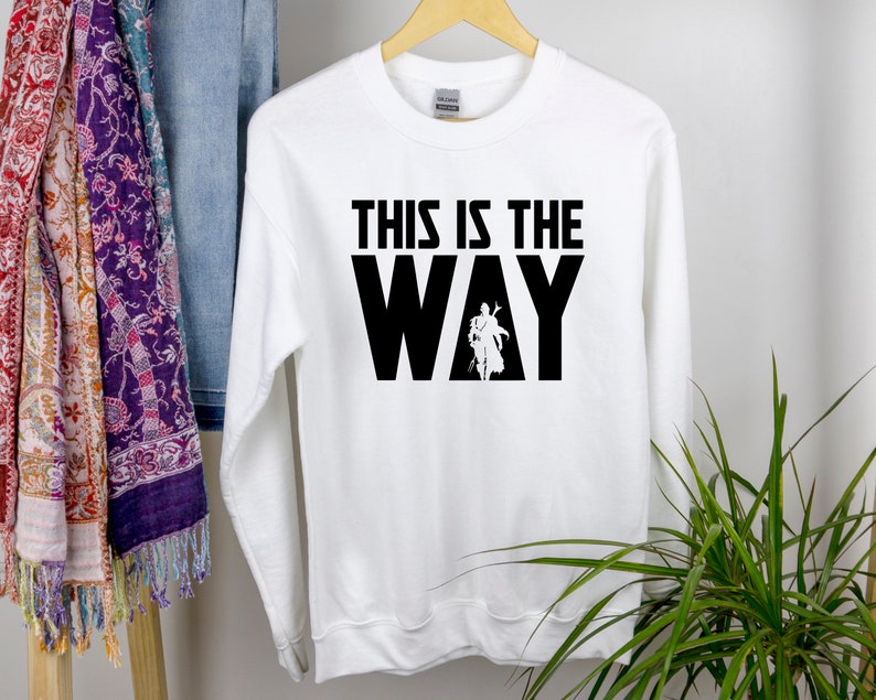 May include: White long-sleeved sweatshirt with black text that reads "This is the Way" and a silhouette of a person in the middle of the word "Way".